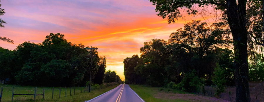 A beautiful sunset centered at the end of a paved country road. The sky is bright yellow at the end of the road, fading to orange and eventually pink as the clouds disperse into the fading blue sky. The road is surrounded by green grass and trees.