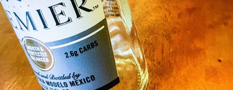 A photo of an empty Corona beer bottle sitting on a wooden table. There is a green book with a red book mark in the background.