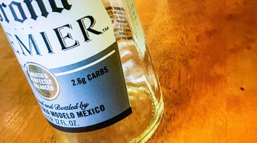 A photo of an empty Corona beer bottle sitting on a wooden table. There is a green book with a red book mark in the background.