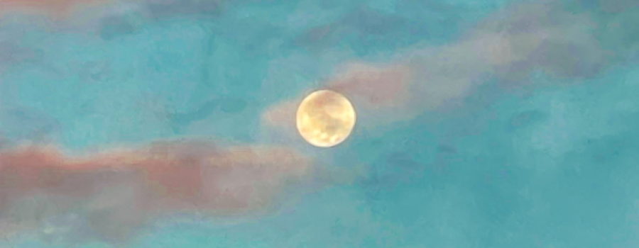 This is a shot of a full moon taken just before dawn. The sky has begun to lighten from black to blue. There are some clouds in the sky which are picking up the pinks of the rising sun. The clouds at the top of the frame are still dark. The moon is centered in the shot and partially covered by clouds.