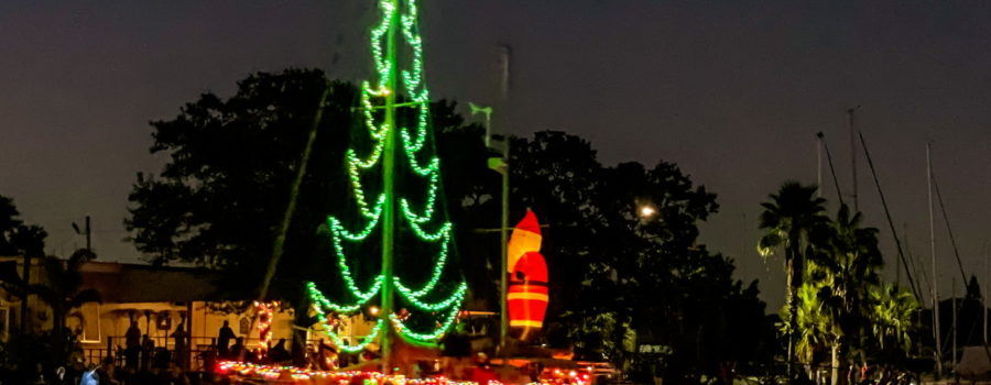 A sailboat cruises down a canal while decorated with Christmas lights including a tree rigged on the mast and a Santa figure on the back.