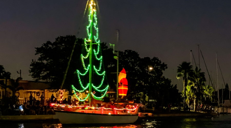 A sailboat cruises down a canal while decorated with Christmas lights including a tree rigged on the mast and a Santa figure on the back.