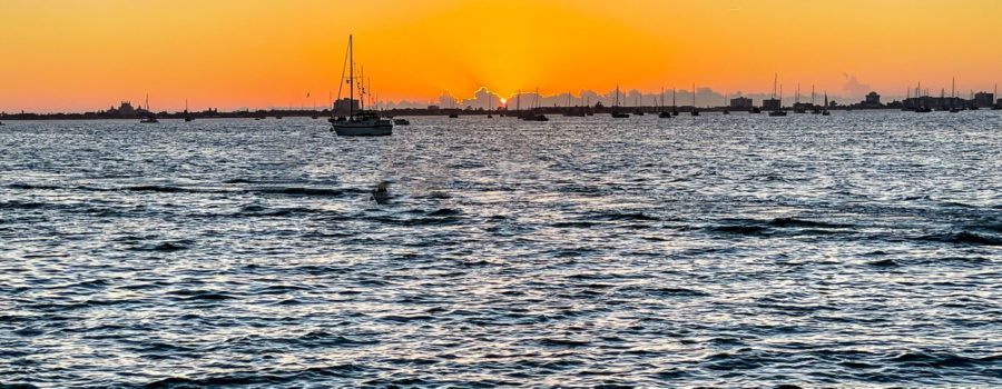 The bright orange ball of the sun can be seen just before going down behind the clouds on the horizon of a boat filled bay.