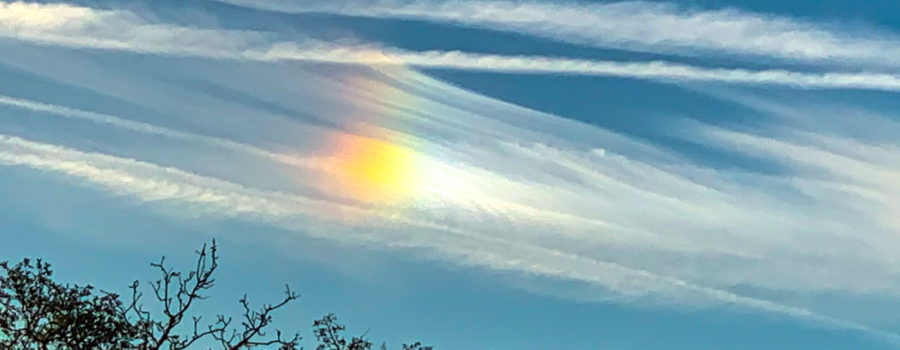 A thin stretch of white clouds reflects the sunlight in just the right way to produce a rainbow of colors, much like a prism does with a light beam.