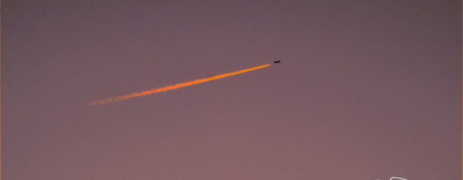 The trail from a large jet airliner lights up an orangish red in the evening sunset, making it look as though the plane is trailing fire.